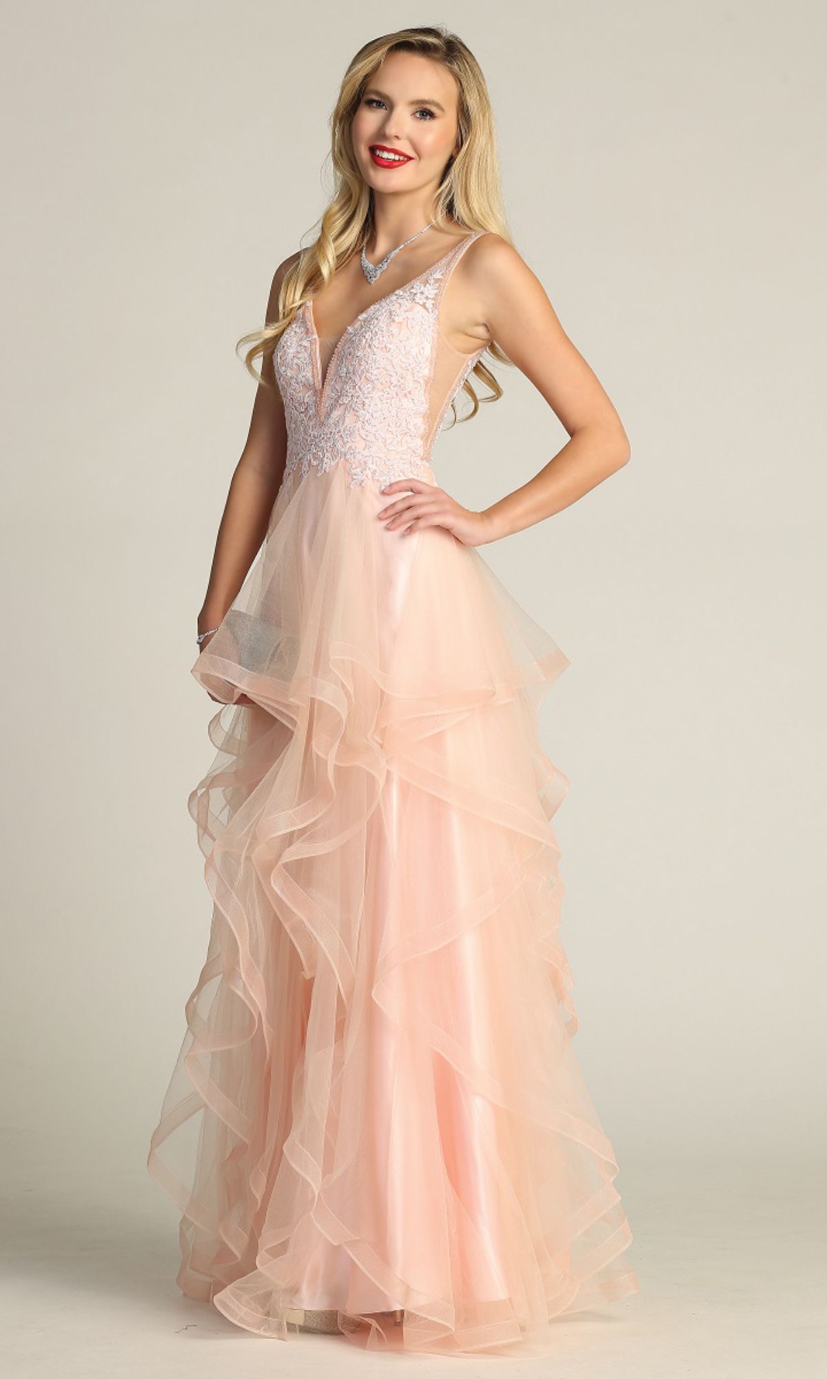 Woman in the Sheer-Sides Long Tiered Prom Dress: Chicas XR6006—light pink, sleeveless, with a lace bodice and layered tulle skirt—stands and smiles against a plain background.